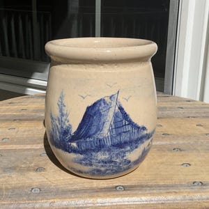 May include: A beige ceramic crock featuring a hand-painted blue scene of a rustic barn, trees, and birds. The vessel has a rounded, wide-mouthed shape with a smooth, speckled finish and a thick, rolled rim at the top edge of the container.