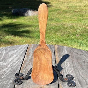 May include: A light brown wooden scoop with a long handle and a wide, flat-bottomed bowl, positioned upright and supported by a black metal stand featuring decorative scrollwork at the base on each side of the wooden utensil.