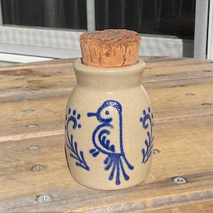 May include: A small, tan ceramic jar with a cork stopper. The jar is decorated with a blue bird design and floral accents. The jar is sitting on a wooden surface.