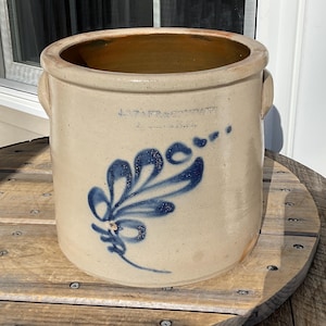 May include: A vintage stoneware crock with a beige exterior and a blue floral design. The crock has two handles and a wide opening. The text "J. & E. FRASER & CO." is visible near the top. It sits on a wooden spool.