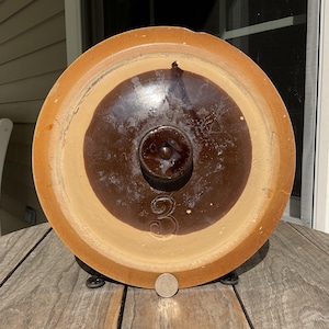 May include: A round, tan stoneware lid with a dark brown glazed center and a central knob handle. The number 3 is embossed on the lower section of the brown glaze. A small silver coin is placed at the bottom edge for scale reference.