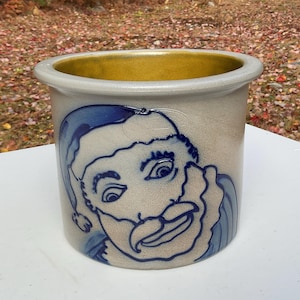 May include: A stoneware crock with a blue Santa Claus face design. The crock has a light gray exterior with a gold-colored interior rim. The Santa Claus face is detailed with blue lines and shading, giving it a vintage look.