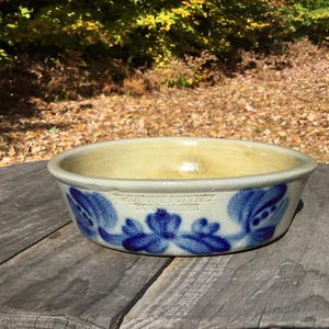 May include: A white ceramic bowl with a blue floral design. The bowl is glazed and has a slightly rounded shape. It is sitting on a wooden surface.