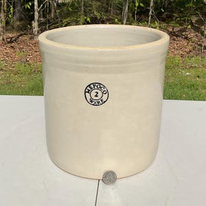 Peut inclure: Un pot en grès de couleur crème avec une large ouverture. Le pot porte un tampon circulaire noir avec l'inscription "MEPCO 2 WARE". Le pot est cylindrique et repose sur une surface blanche. Une pièce de monnaie est au bas du pot.