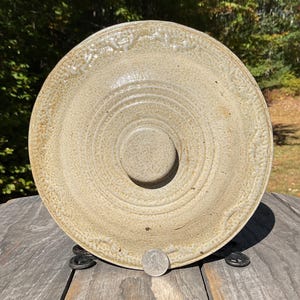 May include: A beige ceramic pottery bowl with a central raised circular feature. The bowl has concentric circular ridges and a textured surface. The rim features a decorative pattern. A quarter coin is visible for scale.