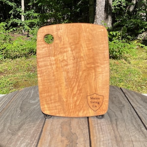 May include: A rectangular light brown wooden cutting board with rounded corners and a circular hole in the top left corner. The board features a natural wood grain pattern and a shield-shaped logo in the bottom right corner that reads Maine Swing Co.