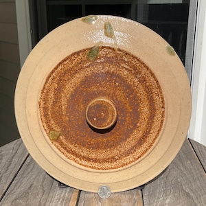 May include: A round ceramic lid with a central knob, featuring a speckled beige outer rim and a textured, dark reddish-brown centre. Three small, raised green leaf-shaped accents are visible on the rim. A US quarter coin is placed at the bottom edge.