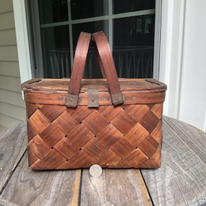 May include: A vintage picnic basket made of woven wood, featuring a rectangular shape with a hinged lid and a dark brown handle. The basket has a metal clasp and is set on a weathered wooden table.