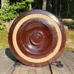 以下が含まれることがあります： 縁に薄いベージュの帯が付いた茶色の陶器の蓋。蓋は光沢仕上げで、円形のデザインです。蓋は金属製のスタンドの上にあり、自然な背景があります。