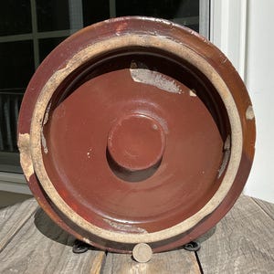 以下が含まれることがあります： 円形のデザインの、大きなアンティークの茶色の陶器の蓋。蓋は中央が盛り上がっており、明るい色の帯が付いた広い縁があります。表面には摩耗や経年劣化の兆候が見られます。木製の表面に硬貨が置かれています。