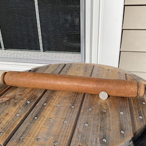 May include: A well-worn, light brown wooden rolling pin, lying on a wooden spool. The rolling pin is cylindrical and shows signs of use. A coin is placed beside the rolling pin for scale.