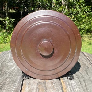 May include: A round, dark brown ceramic lid featuring a smooth, glossy finish with concentric circular ridges and a central knob handle. The lid is positioned upright, showcasing its uniform color and circular design elements on its surface.