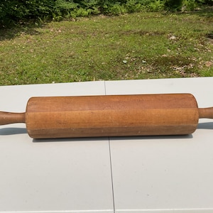 May include: A wooden rolling pin with a cylindrical body and two rounded handles. The rolling pin is made of light brown wood and sits on a white surface. The background features green grass and trees.