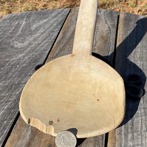 May include: An antique wooden spoon with a broad, shallow bowl and a long handle. The spoon is made of light-coloured wood and shows signs of wear, including a chip on the edge. A quarter coin is placed on the bowl for size comparison.
