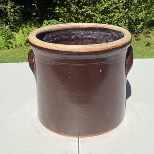Peut inclure: Un pot en grès cylindrique doté d'une glaçure brun foncé très brillante à l'extérieur et à l'intérieur. Le récipient présente un bord épais et arrondi avec une finition beige plus claire et deux anses latérales intégrées pour le soulever.
