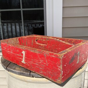 May include: A weathered, rectangular red wooden crate with a handle cut-out. The crate has the number "1" painted in white on the side. The wood shows signs of wear and age, with visible scratches and paint loss. The crate is sitting on a wooden spool.