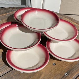 May include: A set of seven white ceramic bowls with a red ombre rim. The bowls are arranged on a wooden surface, showcasing their simple, yet elegant design. The bowls are ideal for serving soups, salads, or desserts.