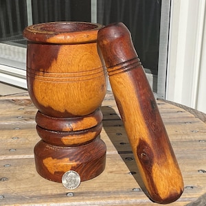 May include: A cylindrical wooden mortar and pestle set featuring a two-toned brown grain pattern. The mortar has a wide, rounded base and a thick rim, while the pestle is long and smooth. A silver US quarter coin is placed at the base for scale.