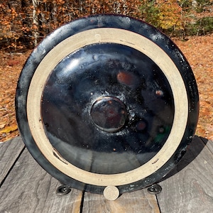 May include: A large, round, black ceramic pot with a lighter-colored rim and a dark, glossy finish. The pot has a circular base and a central, raised knob. The pot is sitting on a wooden surface.