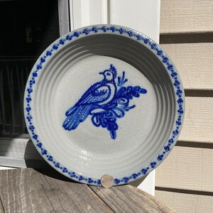 May include: A round, light gray stoneware pie dish featuring a blue hand-painted bird design in the center and a blue floral pattern along the rim. A small silver coin is placed at the bottom edge for scale, showing the dish's relative size.