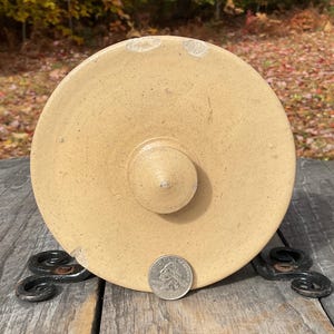 Peut inclure: Couvercle en céramique beige avec un bouton central, probablement pour un pot ou un récipient. Le couvercle est circulaire et a une surface légèrement texturée. Une pièce de monnaie est posée pour comparaison. Le couvercle repose sur un support en métal décoratif.