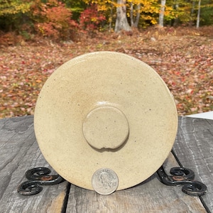 May include: A tan stoneware crock lid with a central handle. The lid is round with a slightly raised edge and a speckled surface. A quarter is placed next to the lid for size reference. The lid is on a decorative metal stand.