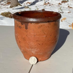May include: A weathered, terracotta-colored ceramic pot with a dark brown rim. The pot has a wide opening and a rounded body, with a small drip of glaze visible on the side. A silver coin rests in front of the pot.