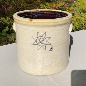 Peut inclure: Un pot en grès cylindrique avec un extérieur crème et un intérieur brun foncé. L'avant présente un motif bleu au pochoir représentant une étoile entourant le chiffre 2. Le récipient comprend deux petites anses latérales et un bord épais.