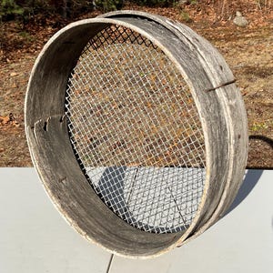 May include: A weathered, round wooden sieve with a metal mesh screen. The sieve's wooden frame shows signs of age and wear, with a natural, gray color. The metal mesh is a fine grid, and the sieve is likely used for sifting flour or other dry ingredients.