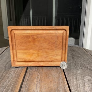 May include: A rectangular wooden cutting board with a smooth, light brown finish and a recessed juice groove running along the perimeter. A silver-colored United States quarter coin is placed at the bottom right corner for scale reference.