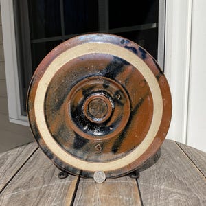 May include: A handmade, ceramic pottery plate with a dark brown, black, and cream color scheme. The plate has a circular design with concentric rings and a central indentation. The number "4" is visible on the plate.