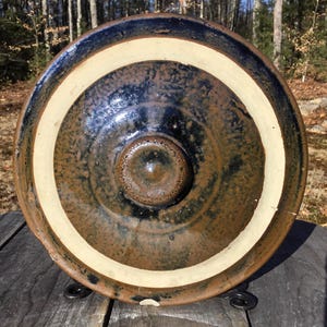 以下が含まれることがあります： 白縁のある茶色と黒の釉薬をかけた陶器の壺の蓋。蓋の中央にノブがあります。