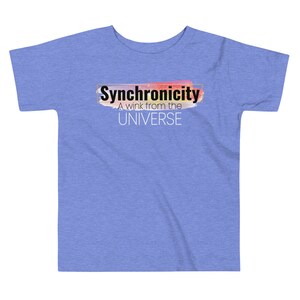 Op de afbeelding: Een lichtblauw T-shirt voor peuters met een kleurrijk abstract ontwerp en de tekst "Synchronicity A wink from the UNIVERSE" in zwart.