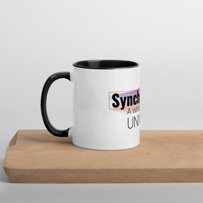 May include: White ceramic mug with a black rim and handle. The mug features a graphic design with the text "Synchronicities" and "A wink from the Universe".