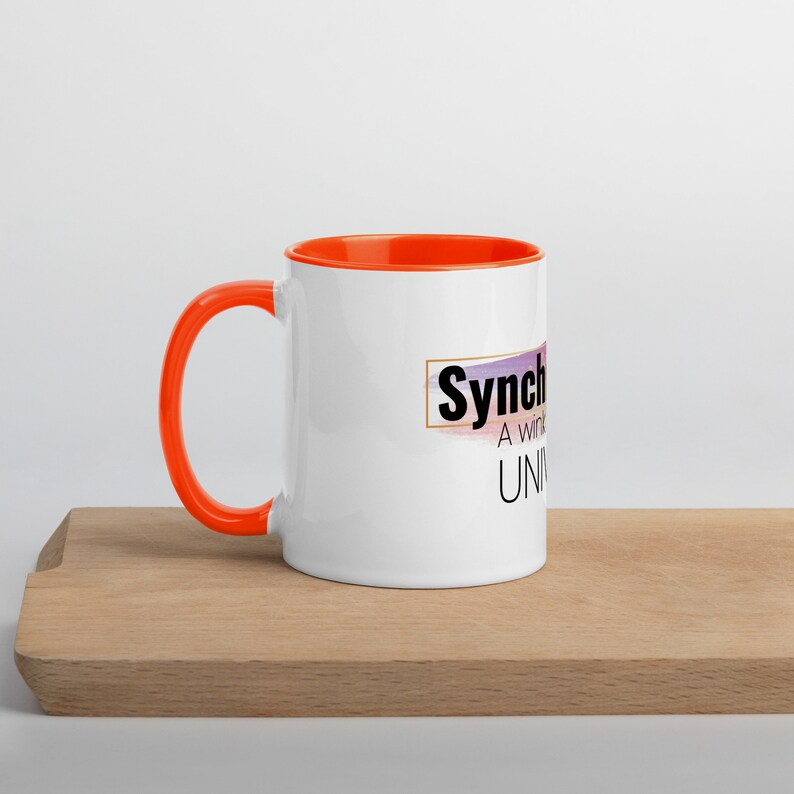 May include: White ceramic mug with an orange rim and handle. The mug features a graphic with the text "Synch A win-win Universe" in a gold rectangle.