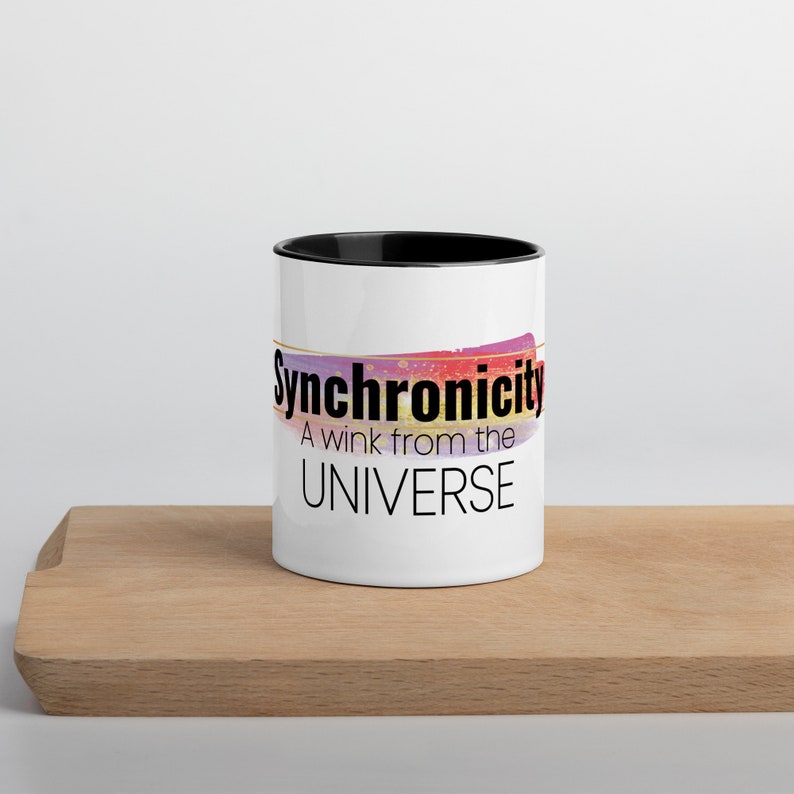 May include: White ceramic mug with a black rim. The mug has a colorful abstract design and the text "Synchronicity A wink from the UNIVERSE".
