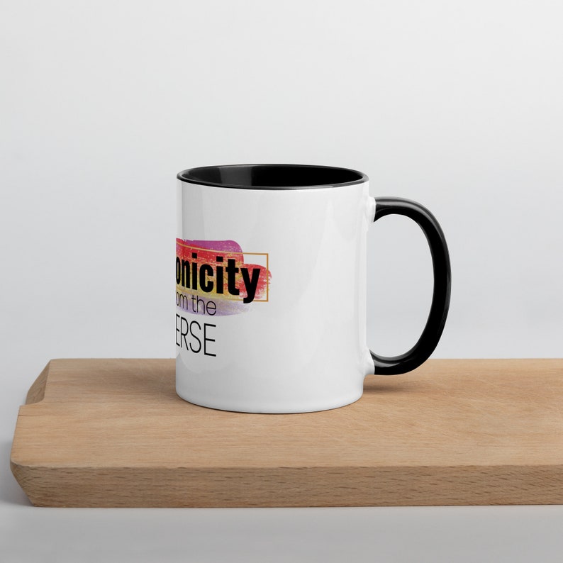 May include: White ceramic mug with a black rim and handle. The mug has a colorful abstract design with the text "Iconicity from the Universe" in black.