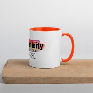 May include: White ceramic mug with an orange rim and handle. The mug features a colorful abstract design with the text "Iconicity from the Universe" in black.