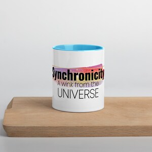 May include: White ceramic mug with a light blue rim. The mug has a colorful abstract design and the text "Synchronicity A wink from the UNIVERSE" in black.