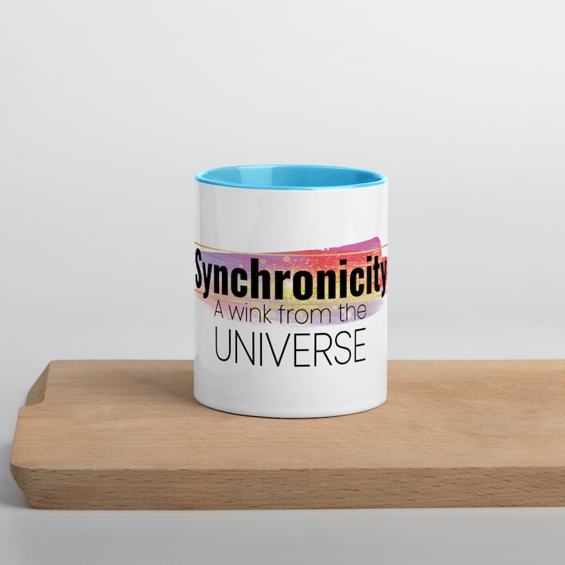 May include: White ceramic mug with a light blue rim. The mug has a colorful abstract design and the text "Synchronicity A wink from the UNIVERSE" in black.