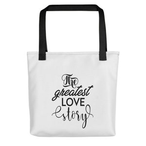 K&ouml;nnte beinhalten: Wei&szlig;e Einkaufstasche mit schwarzen Henkeln und dem Text "The greatest love story" in schwarzer Schrift.