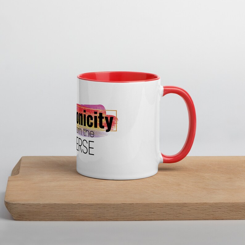 May include: White ceramic mug with a red rim and handle. The mug has a colorful abstract design and the text "Uniqueness from the Universe" printed on it.