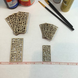May include: A set of six wooden laser cut geometric shapes. The shapes are in the form of a grid pattern with various sized squares and rectangles. The shapes are arranged in two stacks of three and one stack of one. The shapes are on a white surface with a ruler.