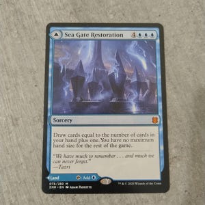 Può includere: Una carta collezionabile Magic: The Gathering Sea Gate Restoration con un'illustrazione blu e bianca di alte strutture geometriche. Il testo include il nome, il costo di mana, il tipo stregoneria, le regole e il testo narrativo di Tazri.