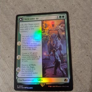 May include: A holographic Magic: The Gathering trading card titled Welcome to... featuring a green border, a T-Rex illustration, and text detailing three saga abilities. The card includes a mana cost of one generic and two green mana symbols.