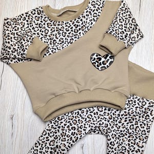Könnte beinhalten: Zweiteiliges Baby-Outfit in Beige und Leopardenmuster. Das Sweatshirt hat Leopardenärmel und ein Herzdetail. Die Leggings und Ärmel haben ein weißes und braunes Leopardenmuster. Das Outfit ist aus weichem Material.