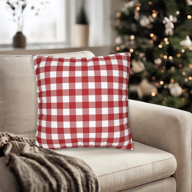 May include: A square decorative pillow with a red and white checkered pattern. The pillow is placed on a beige sofa, with a cozy blanket draped over the arm. A Christmas tree is visible in the background.