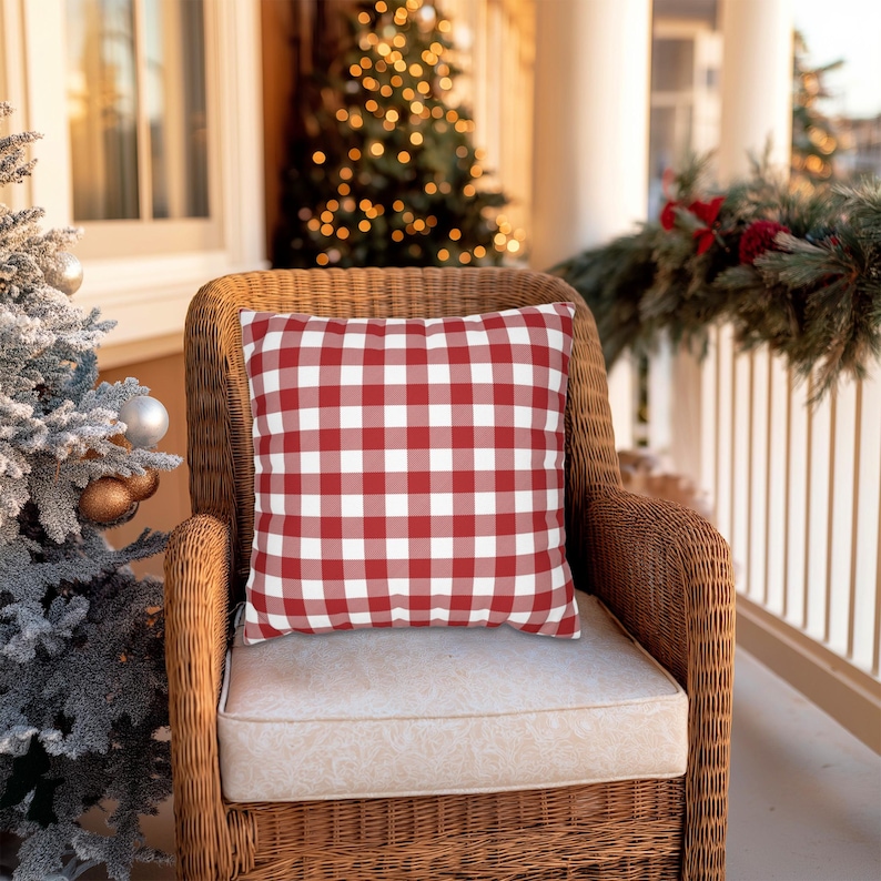 May include: A red and white checkered pillow rests on a wicker chair with a cream-colored cushion. The pillow is the focal point, with a classic gingham pattern. The scene is set on a porch, with a Christmas tree and festive decorations in the background.