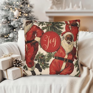 Pu&ograve; includere: Un cuscino bianco con un motivo natalizio festoso con Babbo Natale neri in costume rosso e rami di agrifoglio verdi. Il cuscino ha un cerchio rosso con la parola "Joy" in caratteri bianchi.