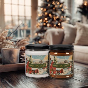 May include: Two glass jar candles with black lids. One candle is white, the other is brown. Each jar has a label with a winter scene of a red house and the text "peace on earth". The candles are on a wooden surface.